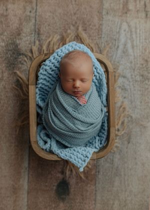 Sleeping newborn baby swaddled in blue wrap resting in a wooden bowl on rustic floor.
