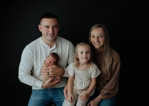 Happy family of four posing against black backdrop with newborn baby and young toddler girl in studio.