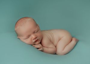 Newborn baby sleeping curled up on a teal blue background in a classic posed newborn portrait.