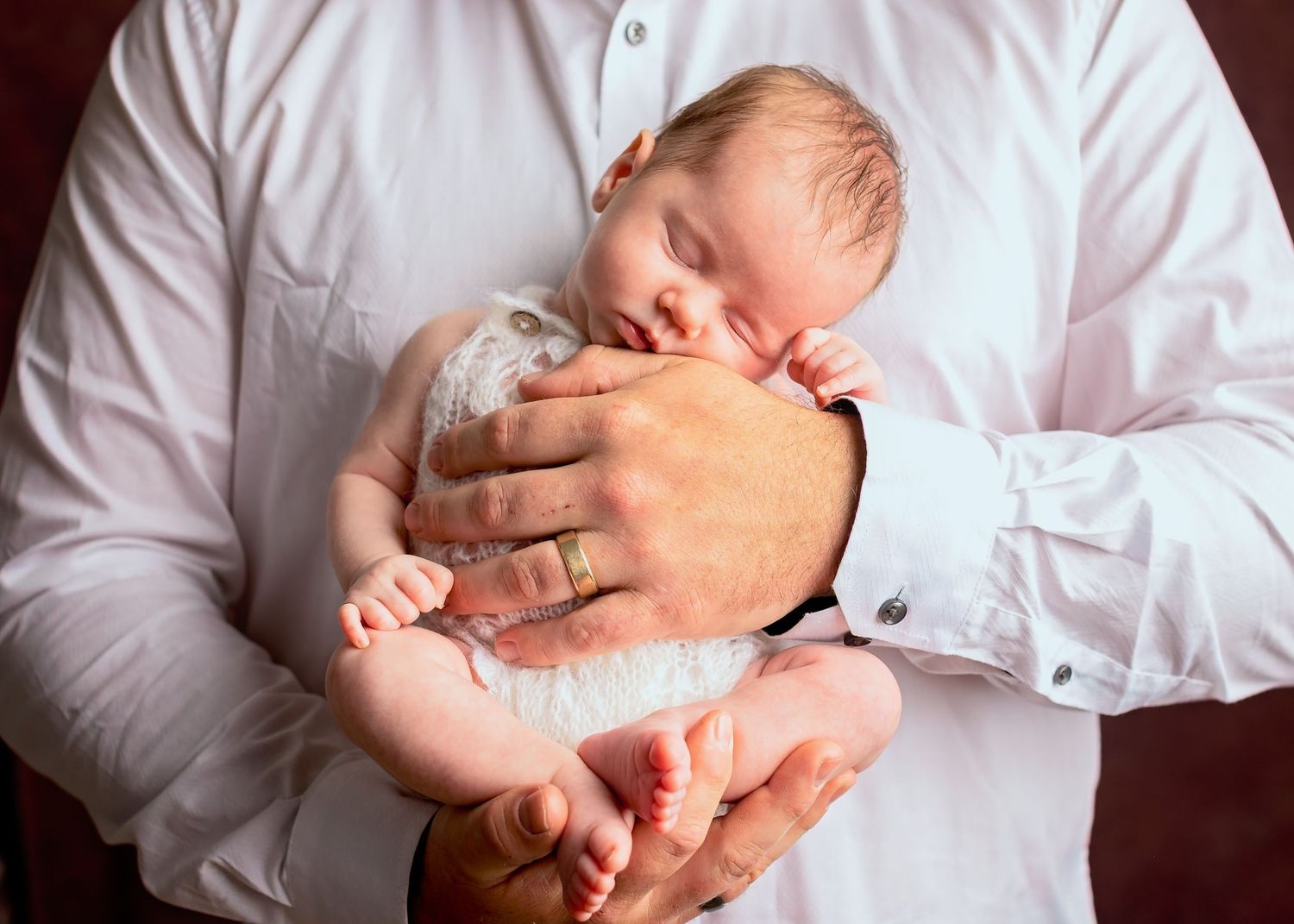 A newborn baby sleeps peacefully while being cradled in gentle hands wearing a white shirt.