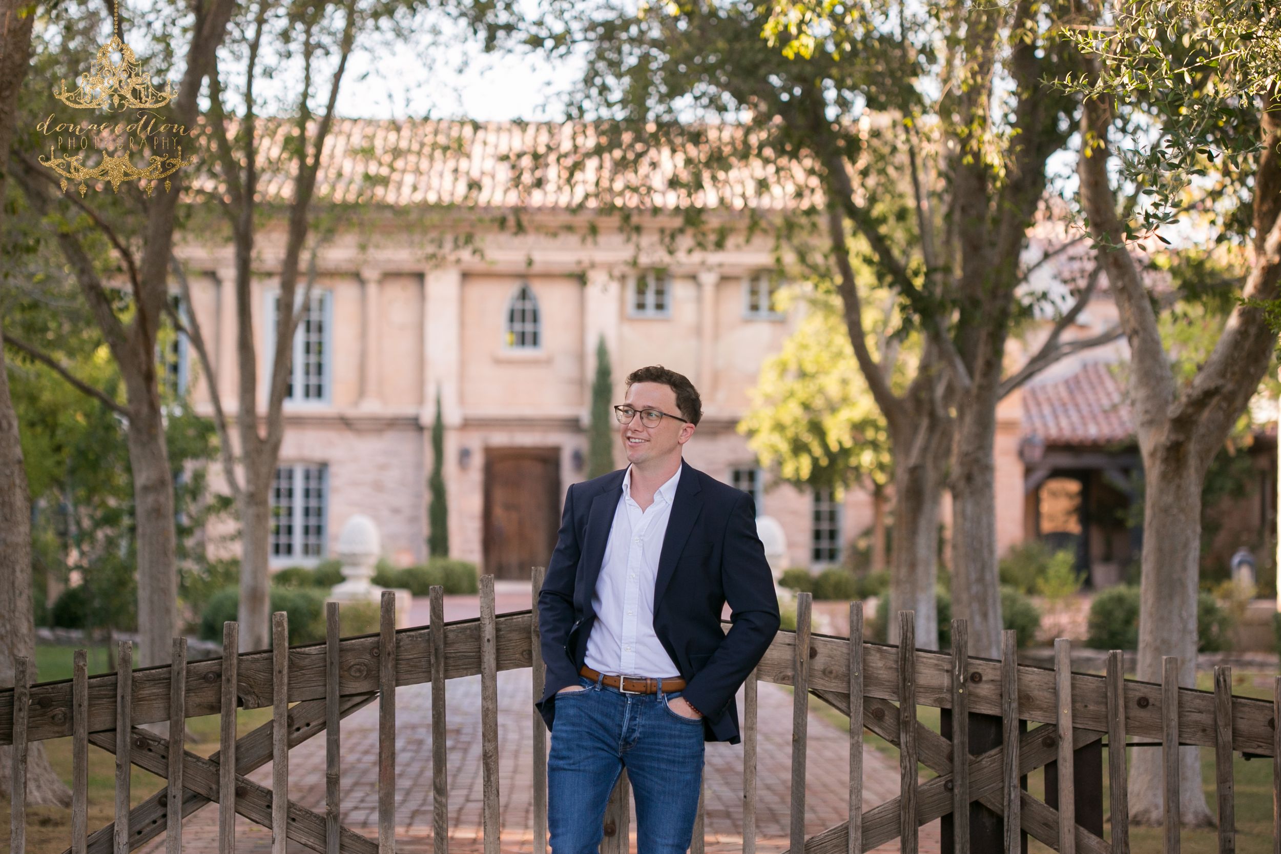 Smiling man in navy blazer and jeans standing on a wooden bridge outside an elegant building.
