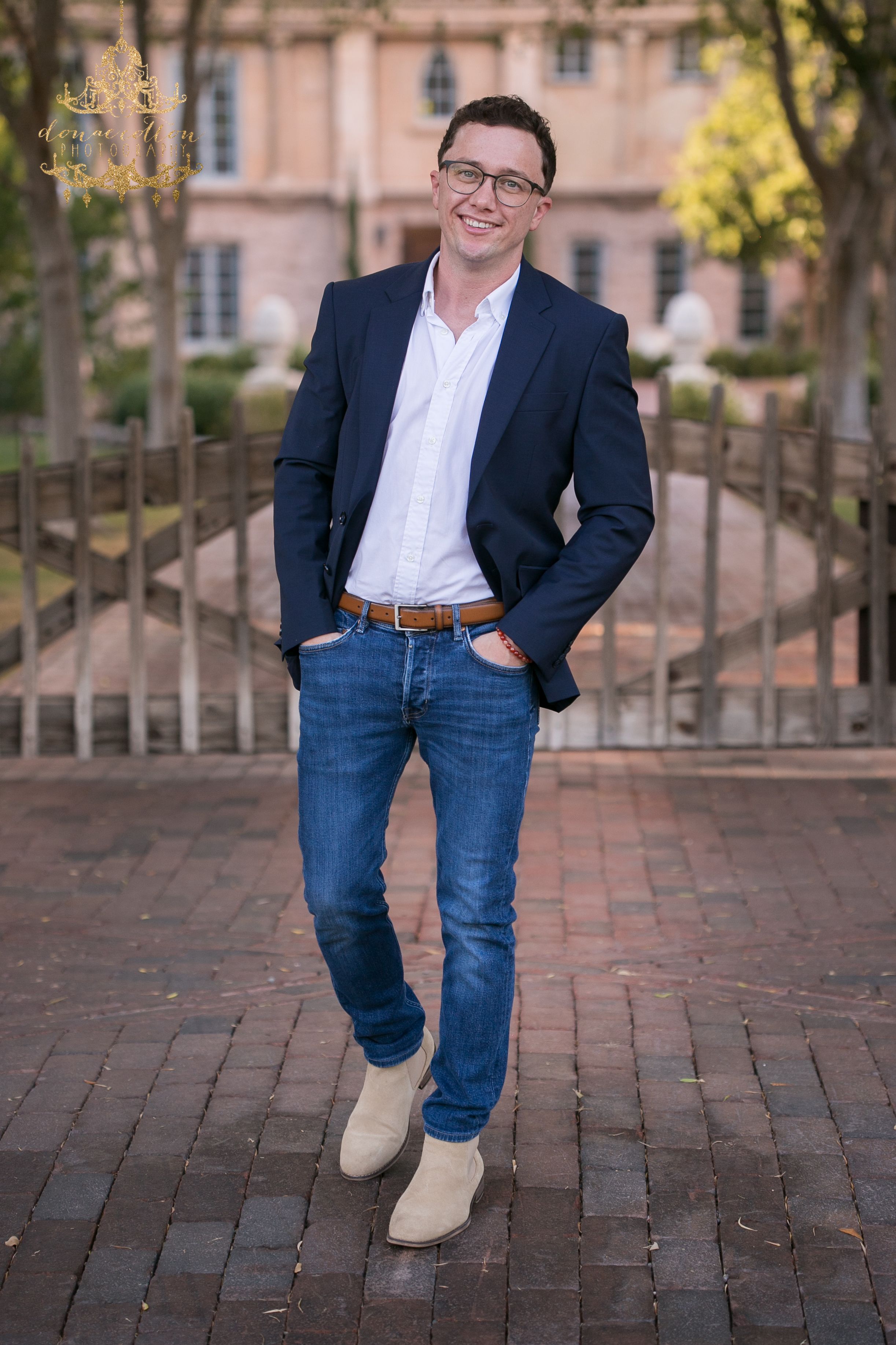 Young man in navy blazer, jeans, and tan boots smiling on a brick pathway near a building.