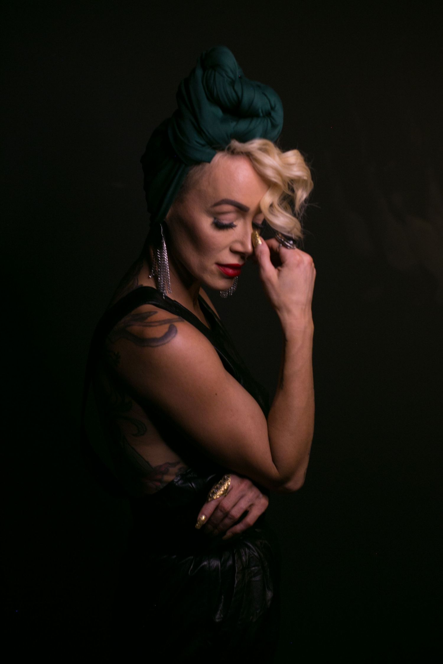 Blonde woman with green turban in black leather outfit posing with eyes closed in dark studio.