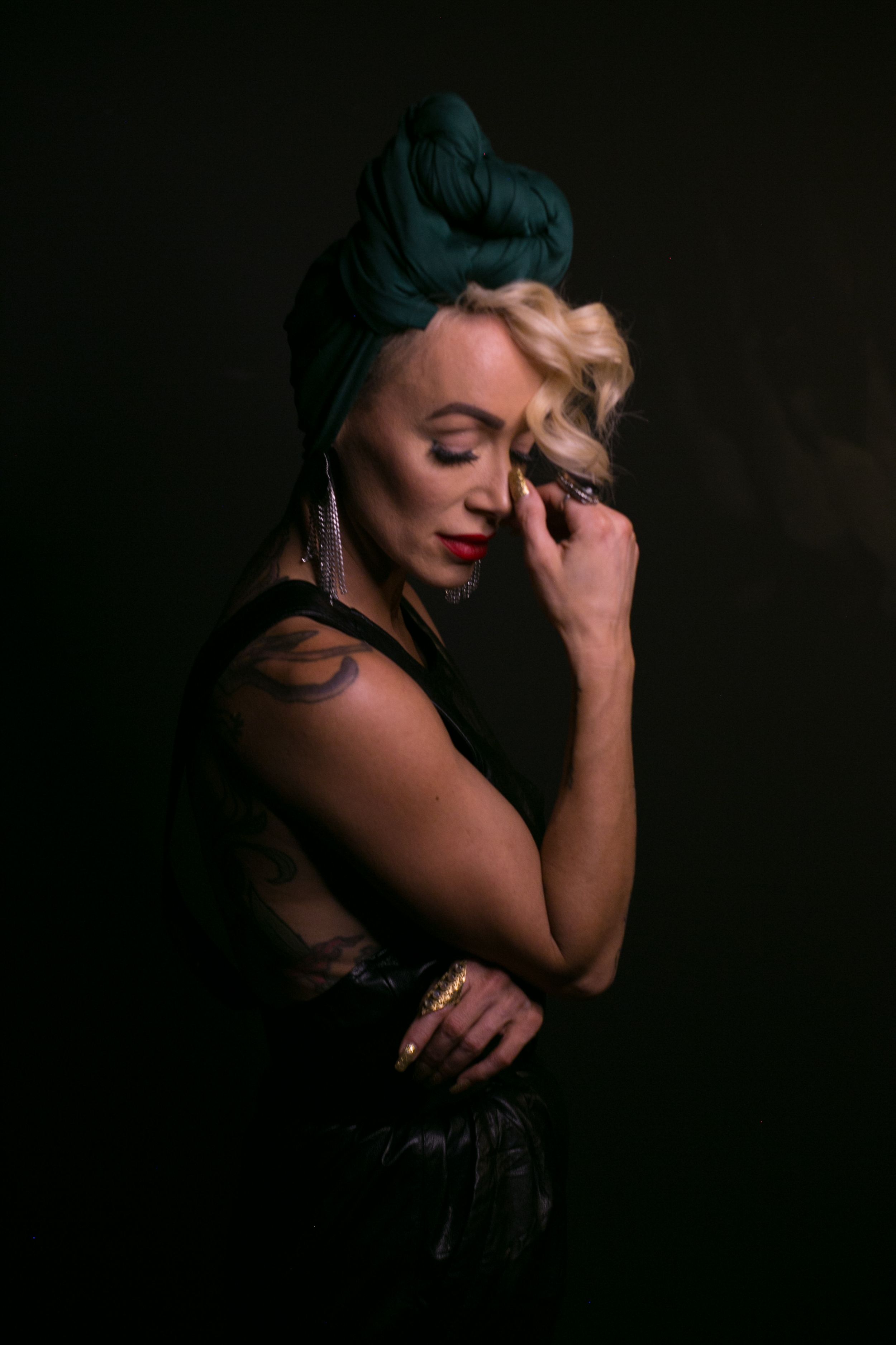 Blonde woman with green turban in black leather outfit posing with eyes closed in dark studio.