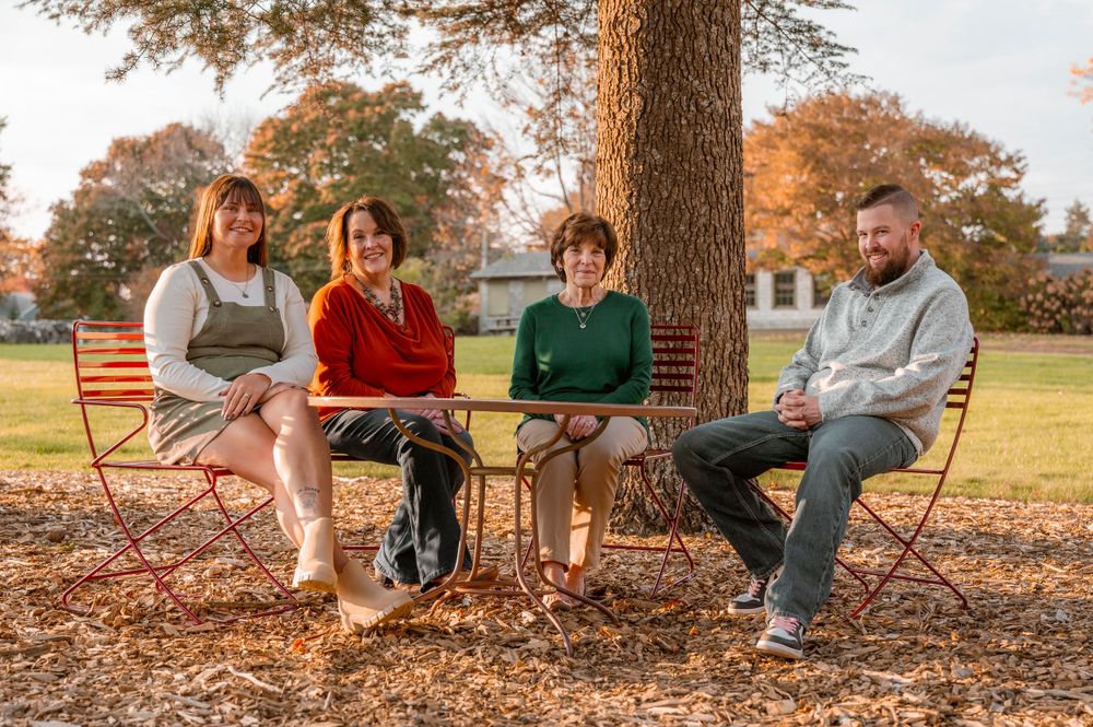 Four people sit casually on wooden chairs under a large tree during autumn.