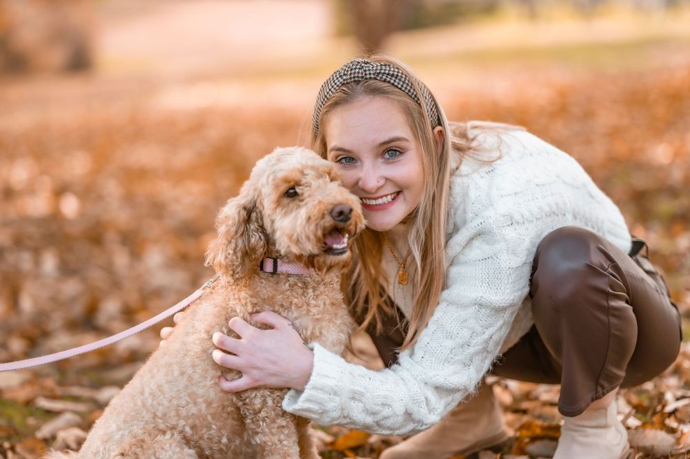 Person in white sweater hugging goldendoodle dog among fallen autumn leaves.