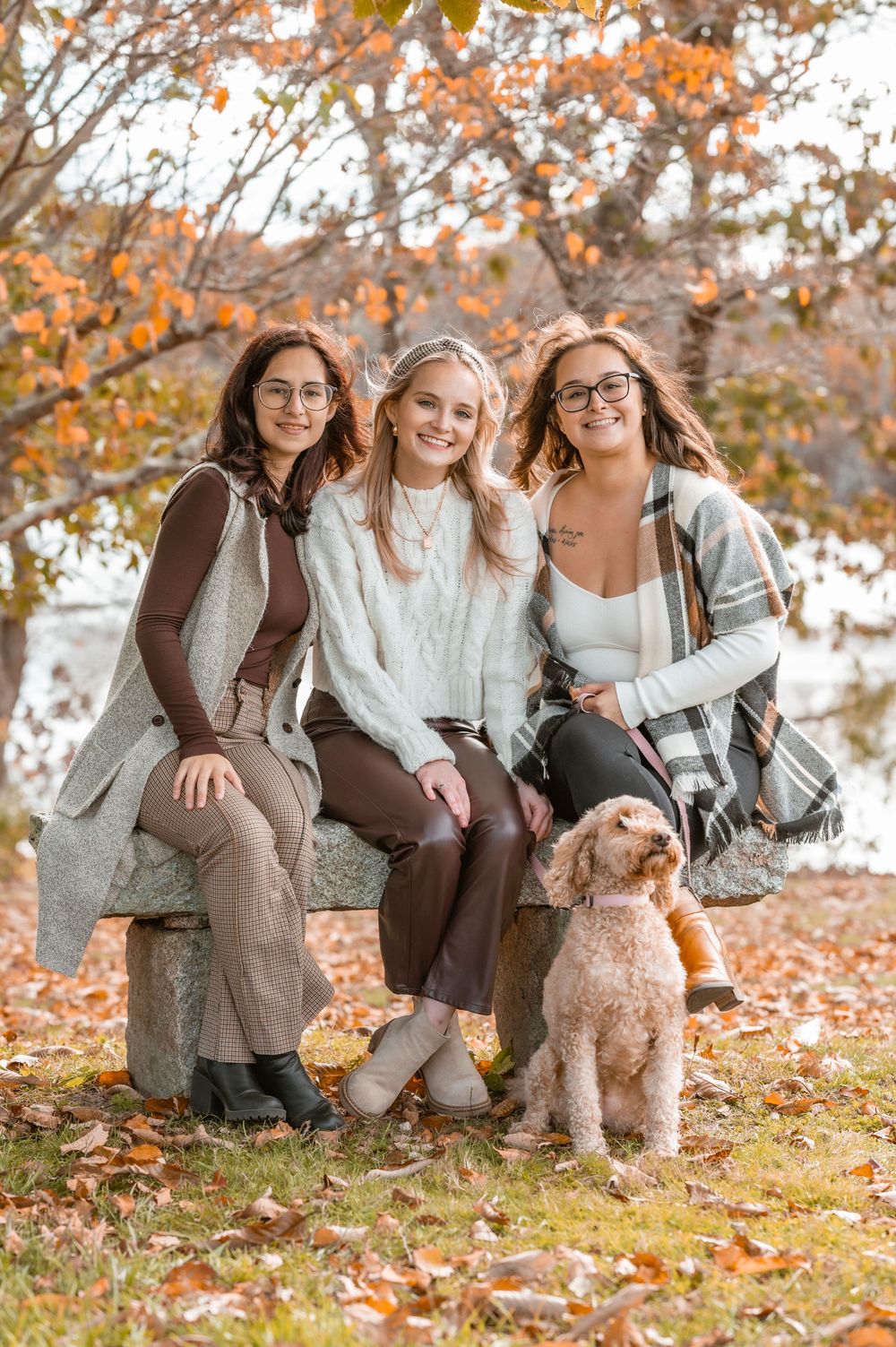 Three friends sitting on bench with goldendoodle dog during fall season.