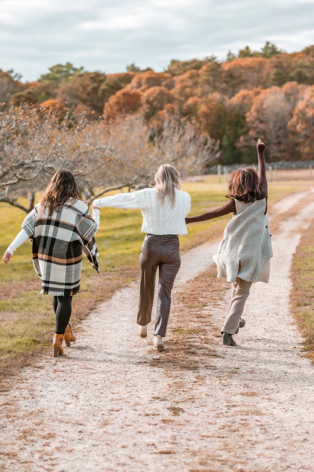 Three friends run joyfully down an autumn path wearing cozy sweaters and casual attire.