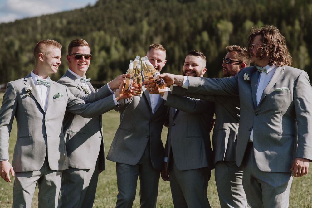 A group of groomsmen in gray suits toast with beer glasses against a mountain backdrop.