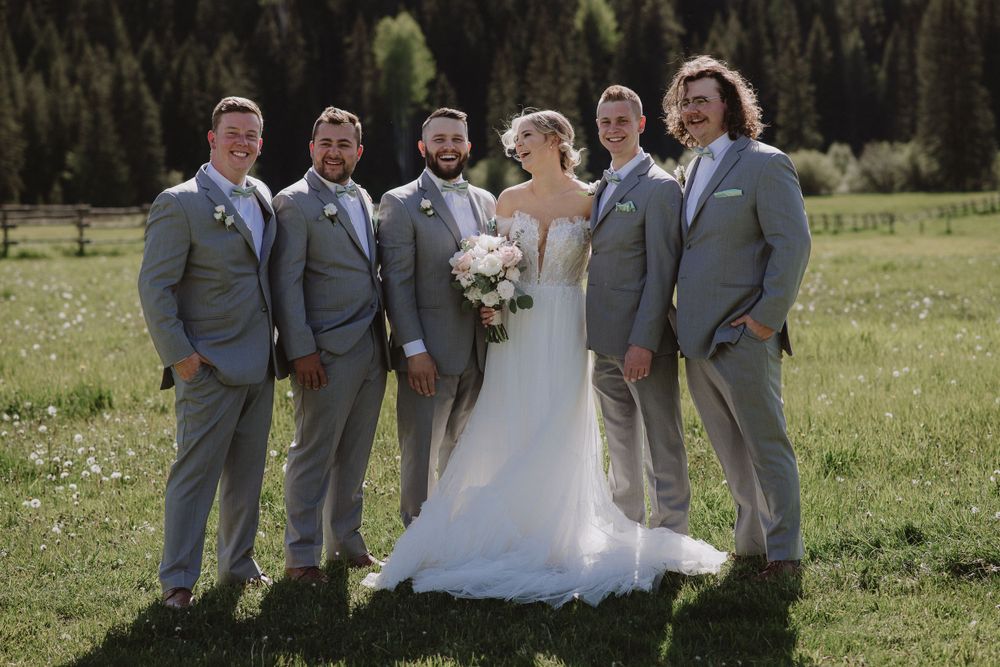 Wedding party in grey suits posing together in a green meadow with evergreen forest backdrop on a sunny day.