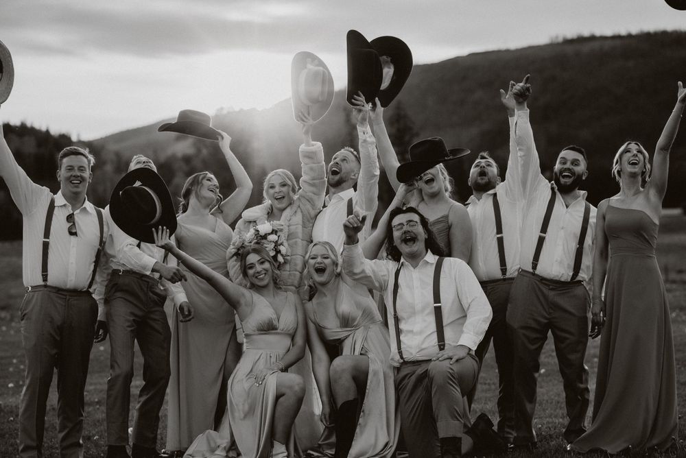A joyful wedding party celebrates outdoors with raised arms and hats against a sunlit mountain backdrop.