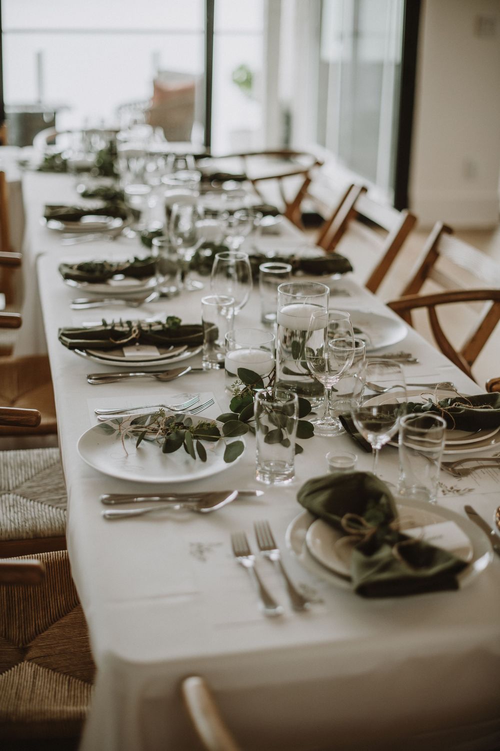 An elegant table setting with white linens, rustic wooden chairs, and fresh eucalyptus garland running down the center.
