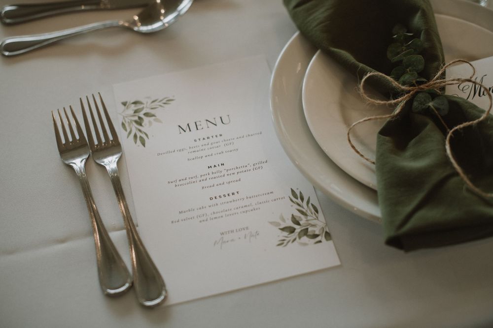 Elegant wedding menu card with green floral design placed beside silver forks and a green napkin on white tablecloth.