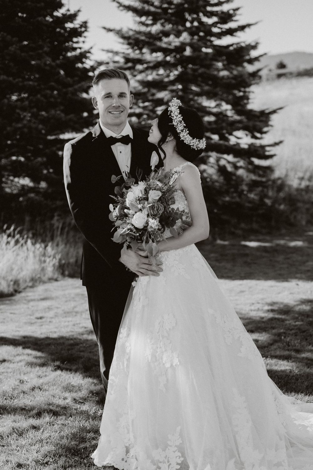 Black and white wedding photo of couple in formal attire with evergreen trees in background.