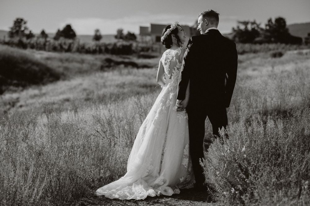 A bride and groom stand together in a field of tall grass at sunset, captured in black and white photography.