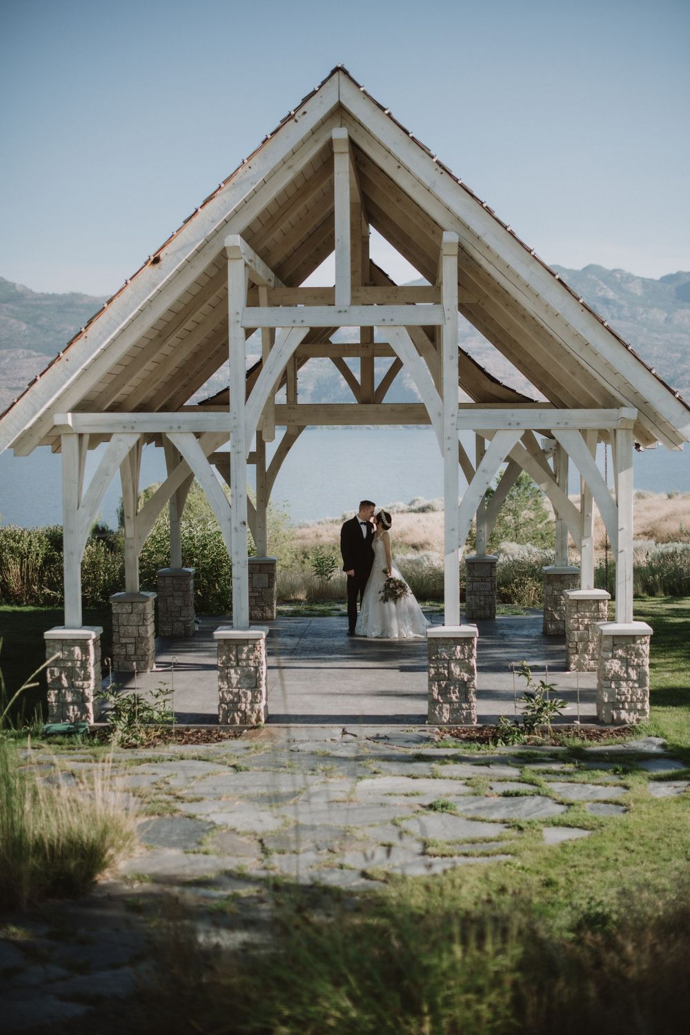 Elegant white timber-frame wedding pavilion with stone pillars set against mountain landscape and blue sky.
