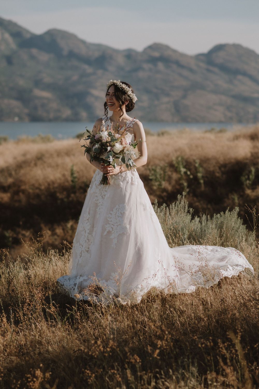 A bride in a lace wedding gown holds a bouquet while standing in a field with mountains in the background at sunset.