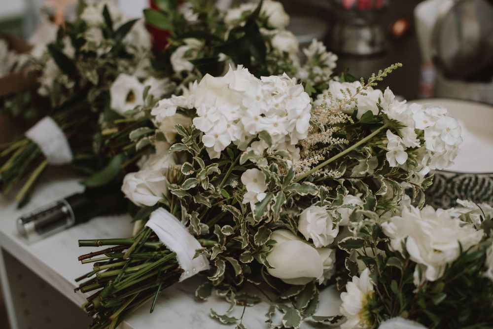 A delicate wedding bouquet with white roses, hydrangeas and mixed greenery tied with white ribbon rests on a table.