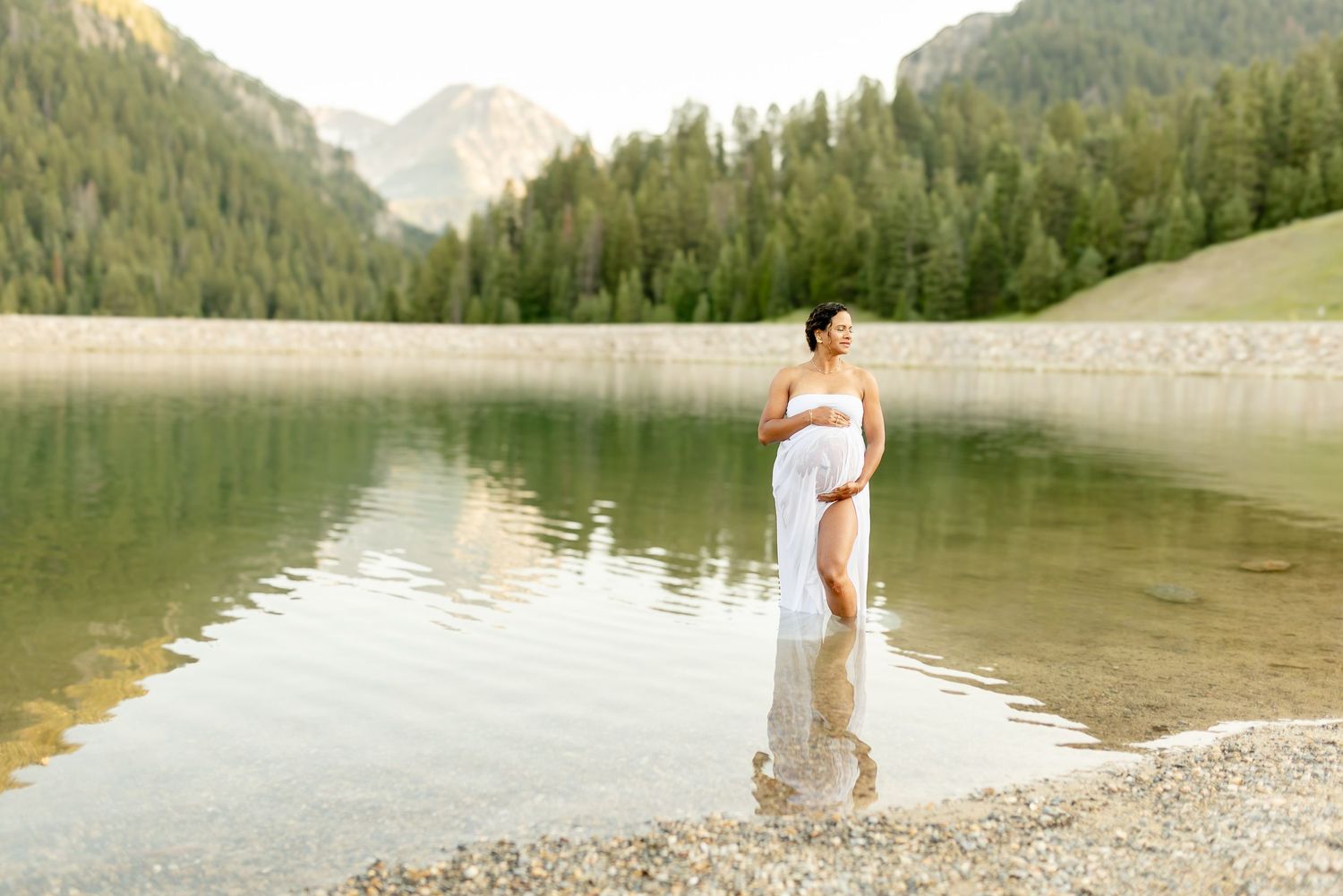 Elegant figure in black gown poses dramatically on mountain road with snow-capped peaks and evergreen forest backdrop.