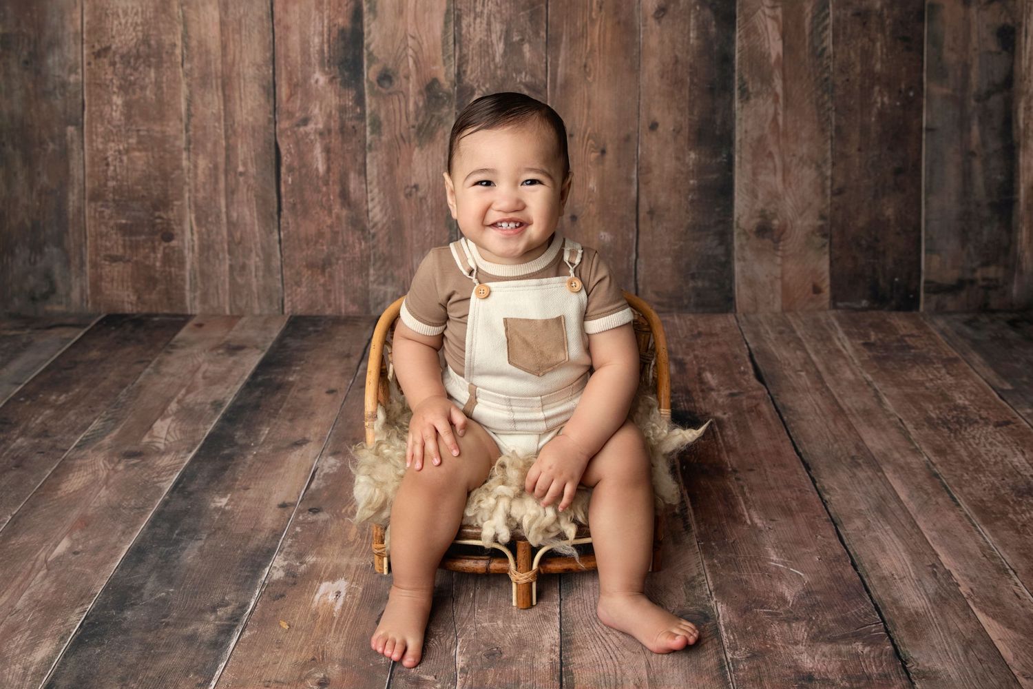 Baby in vintage overalls sits on wooden tricycle against rustic backdrop.