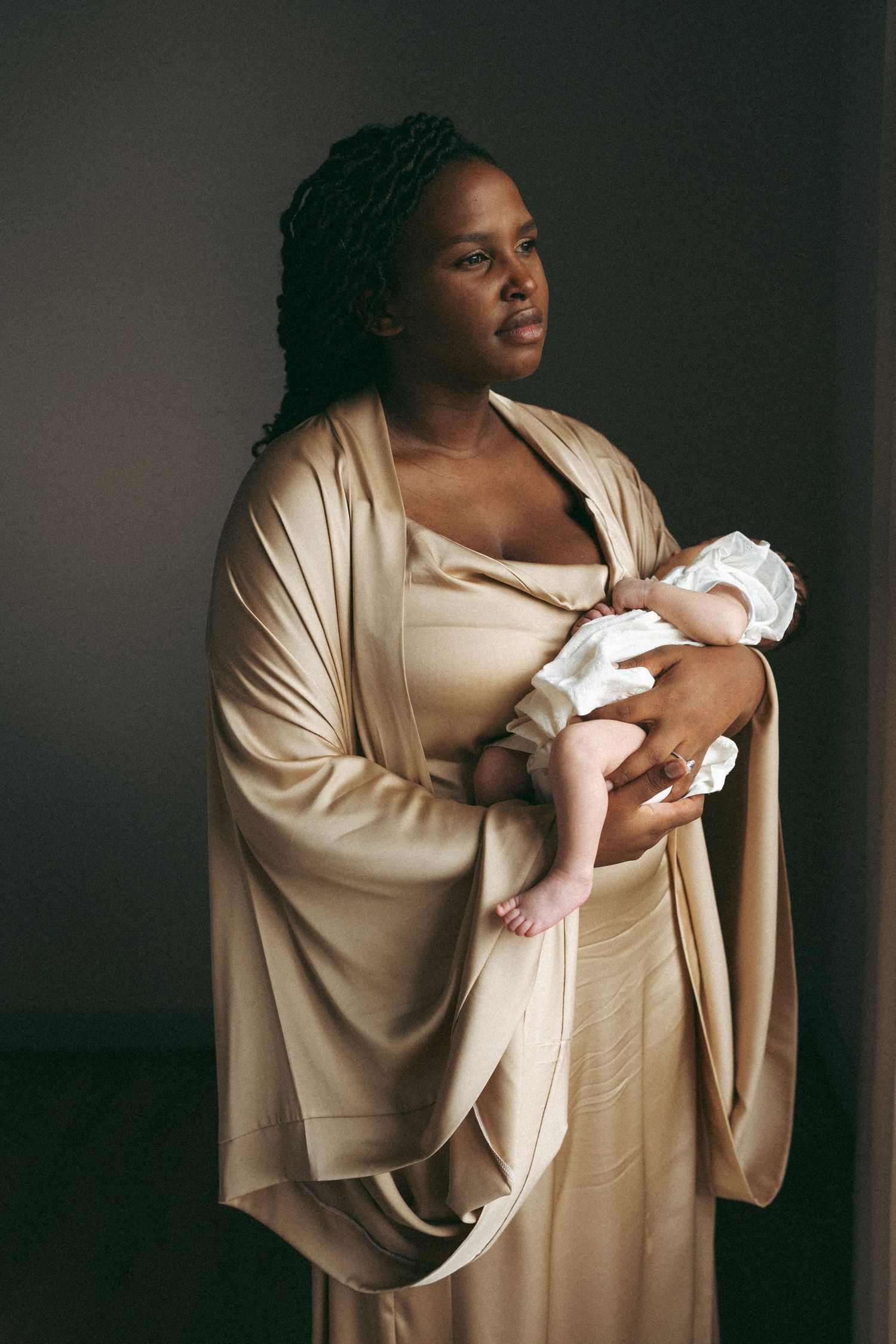 Dramatic portrait in beige clothing holding newborn against dark background.