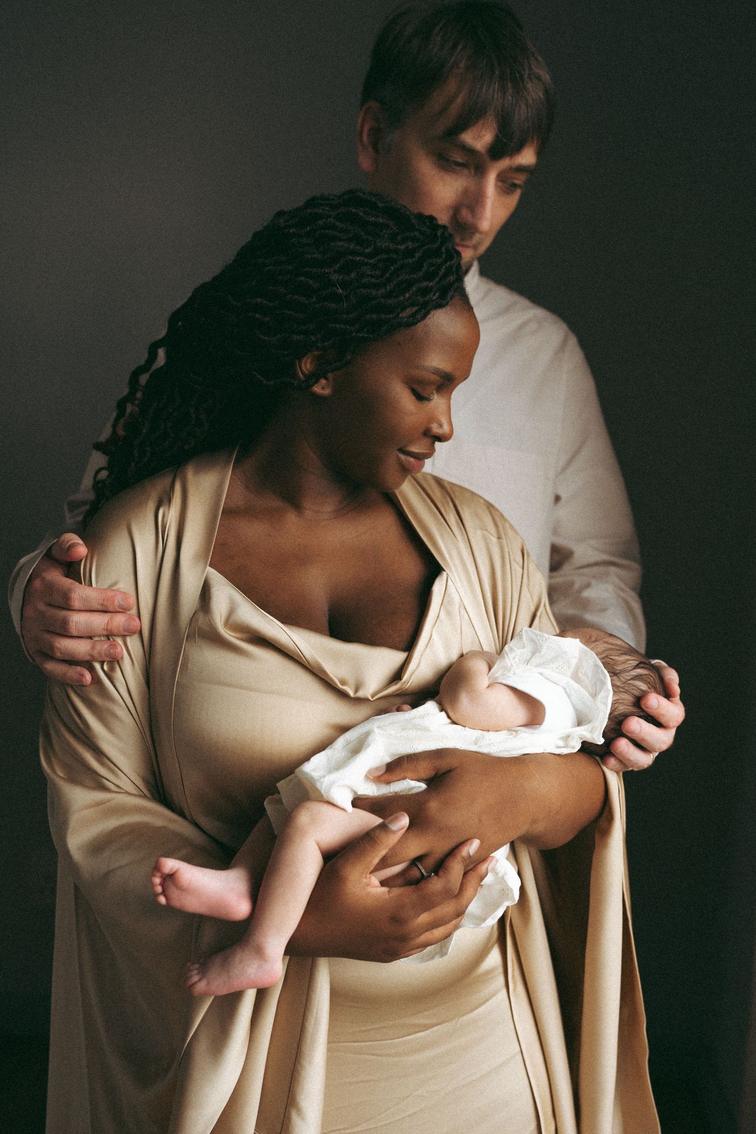 Parents in light colored clothing share an intimate moment while holding their sleeping newborn baby.