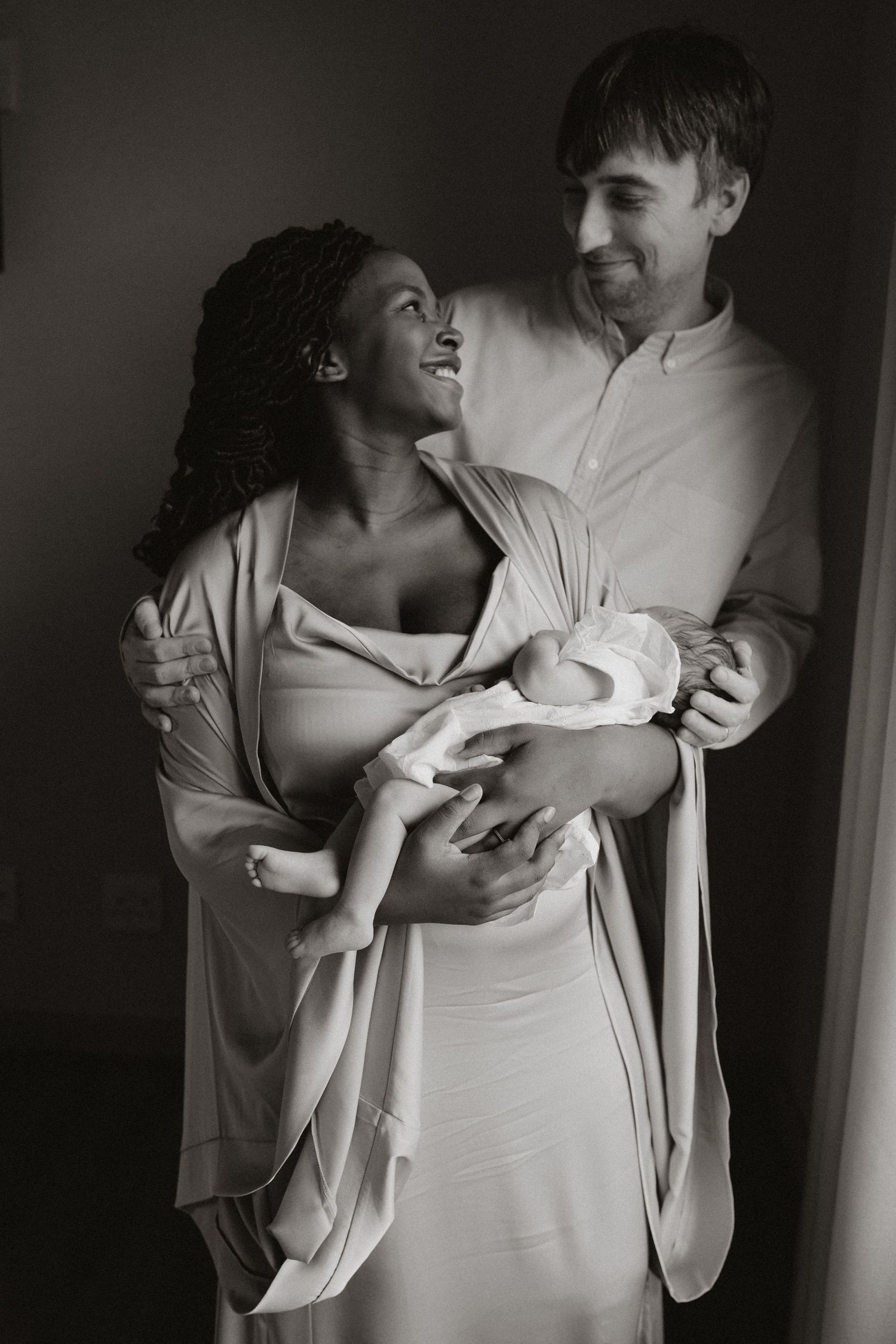 A couple gazes lovingly at their newborn baby while wearing cream colored clothing in a dark studio setting.