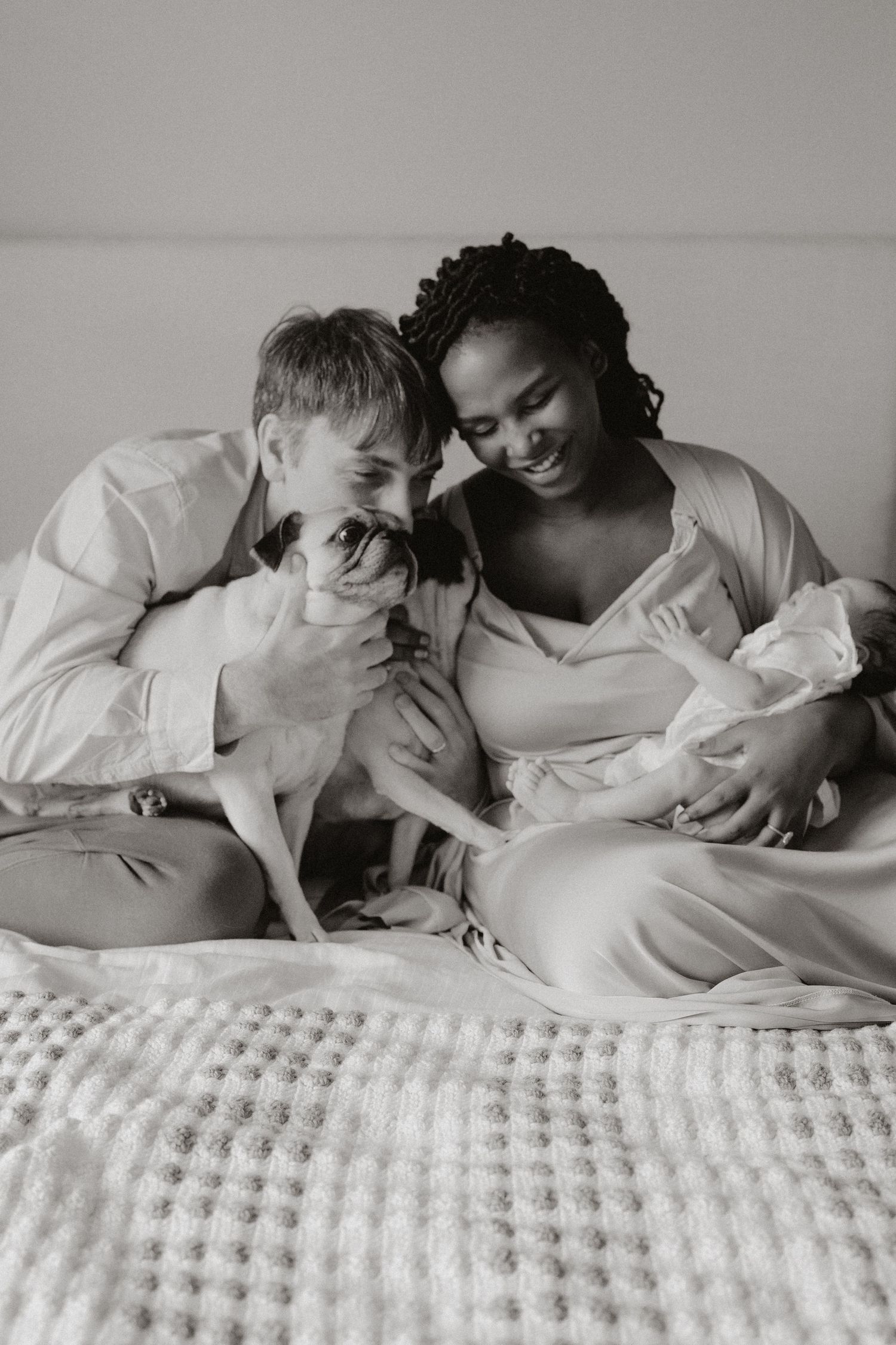 Happy couple lounging on bed with their dog and newborn baby in black and white photography.