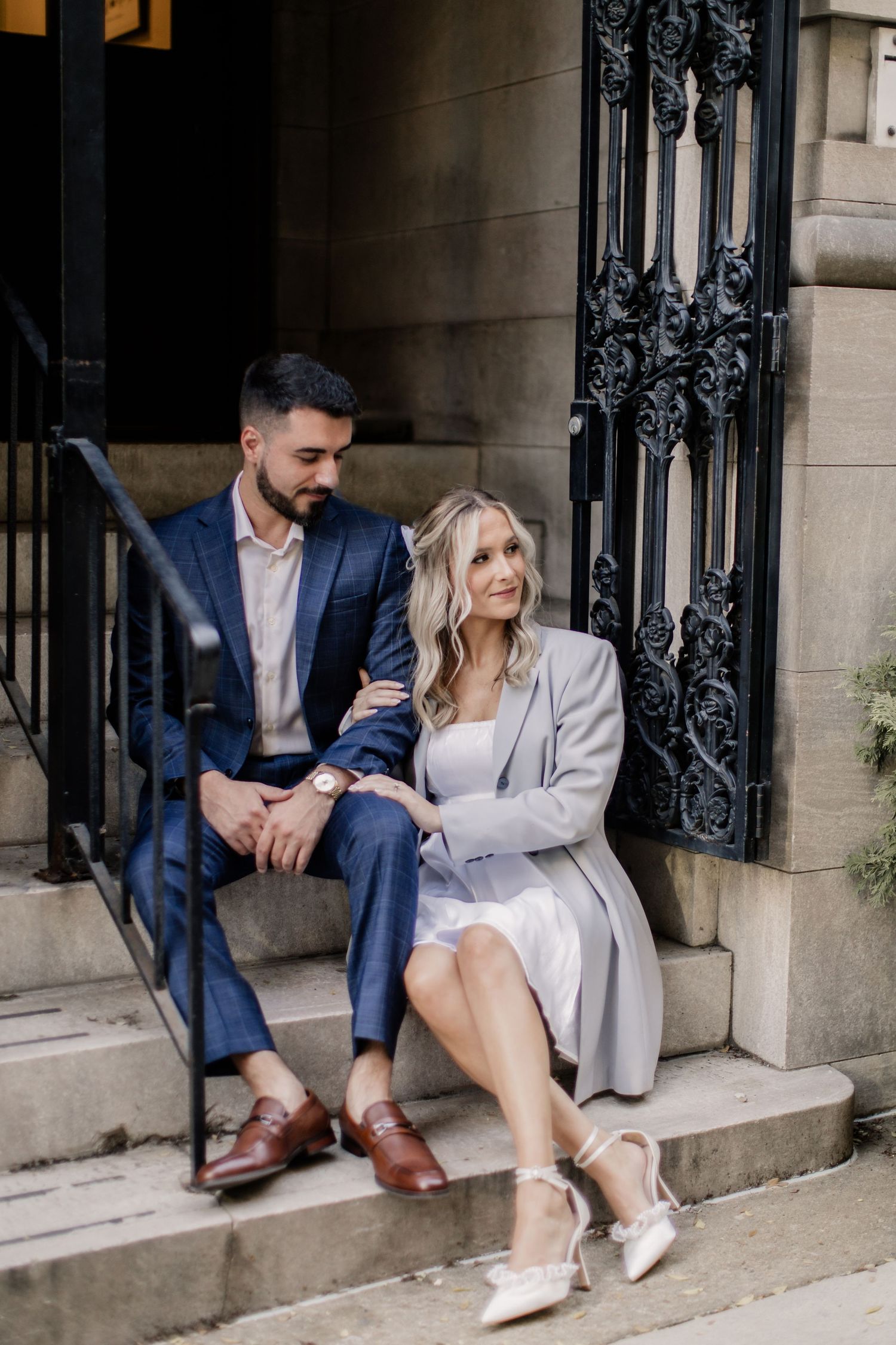 A couple in formal attire sits casually on stone steps outside an elegant building with ornate iron gates.