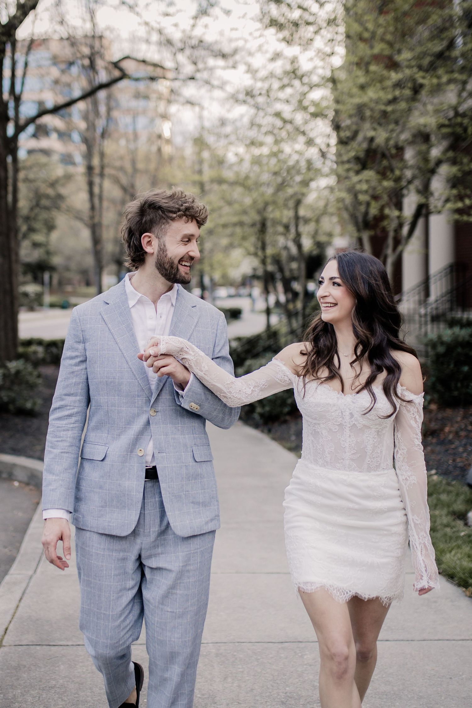 A couple in wedding attire shares playful moments while walking along a tree-lined path in a romantic photoshoot series.