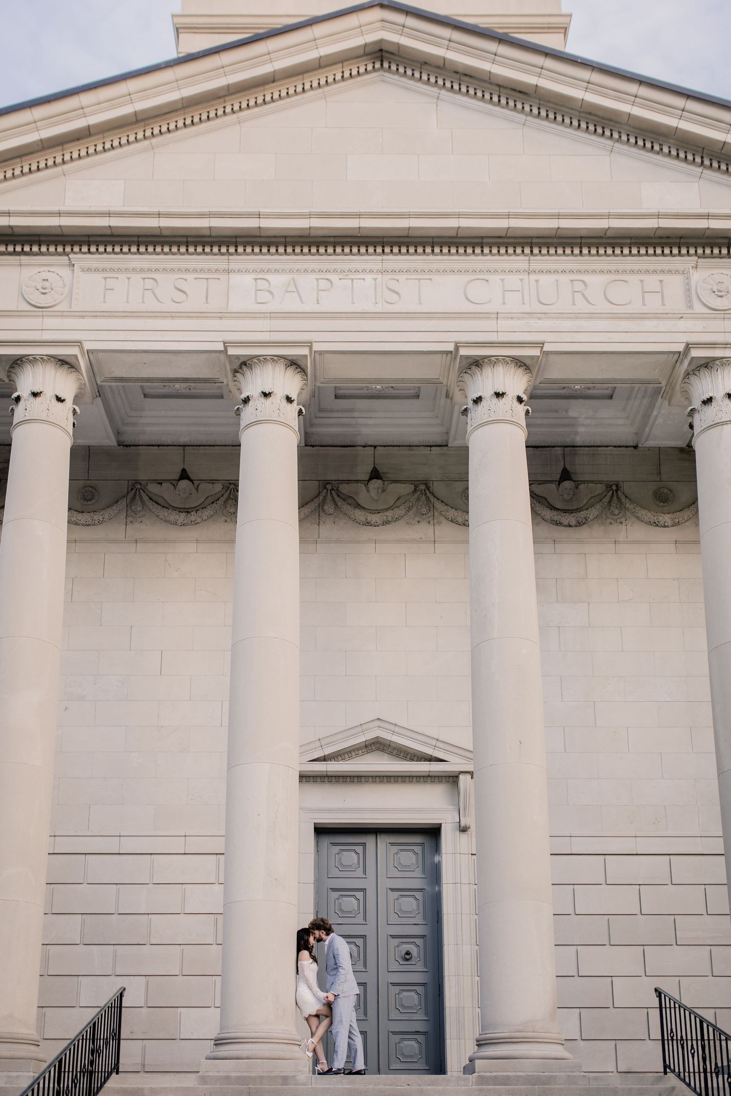 Neoclassical First Baptist Church facade with white stone columns and blue doors in a panoramic view.