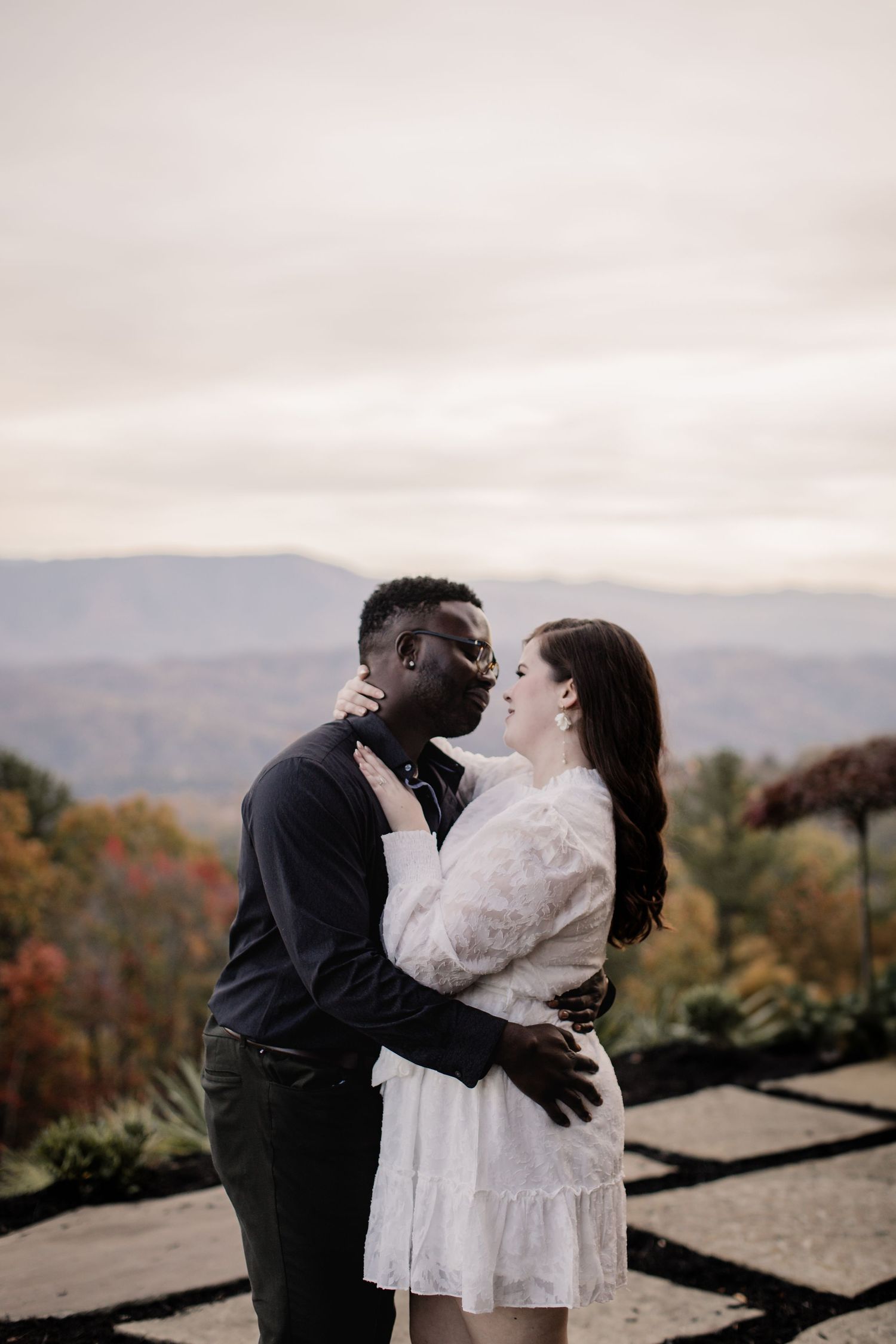 A romantic couple embraces on a mountain overlook with autumn foliage and misty hills in the background.