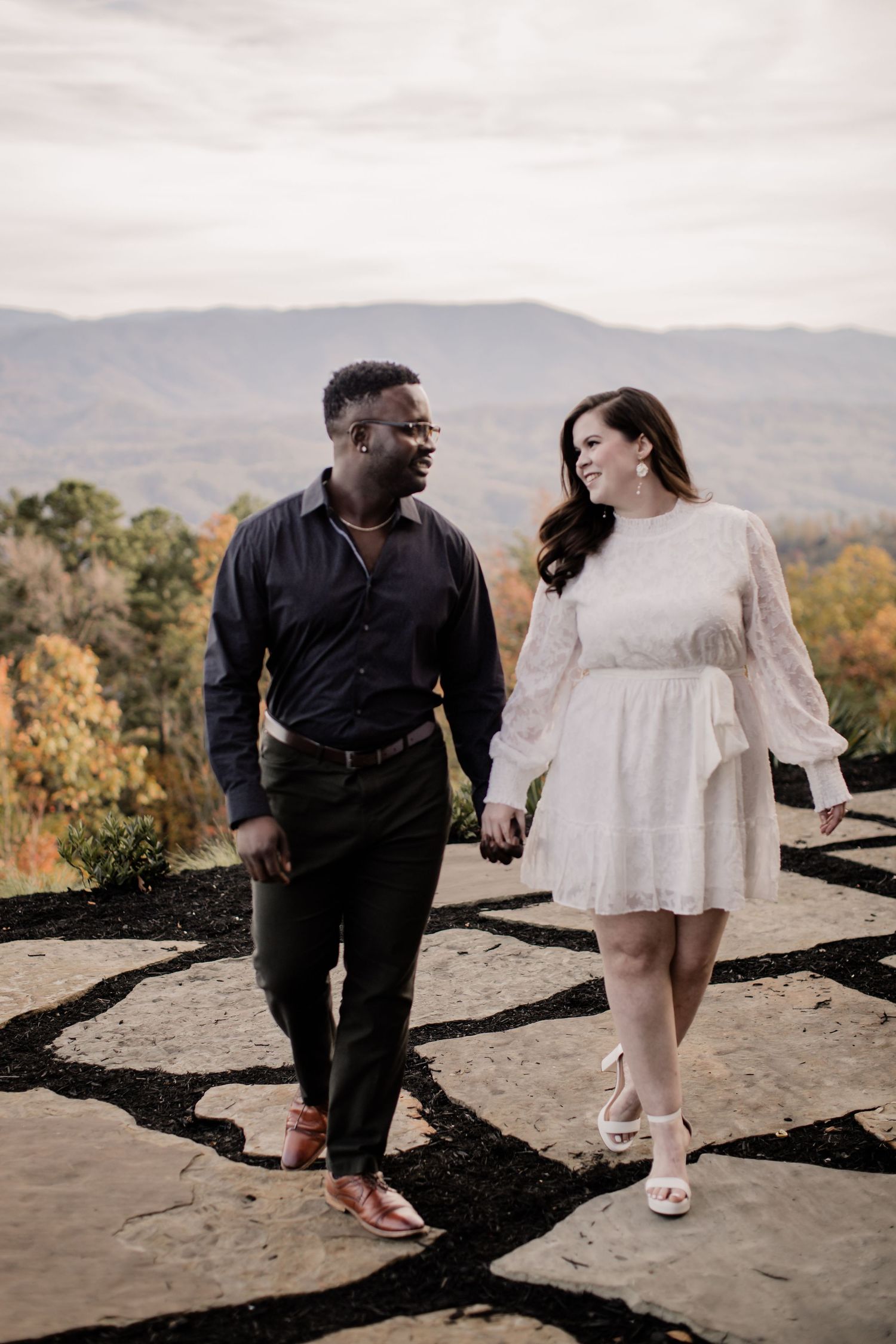 A couple in contrasting black and white outfits holds hands while walking along a scenic mountain overlook at sunset.