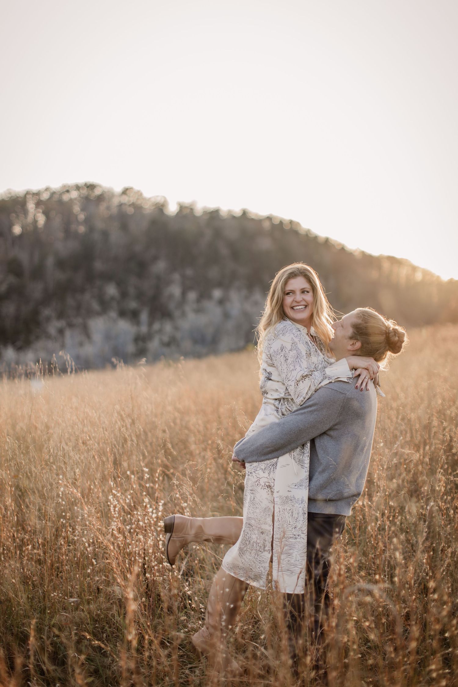 A couple embraces in a golden field at sunset with mountains in the background.