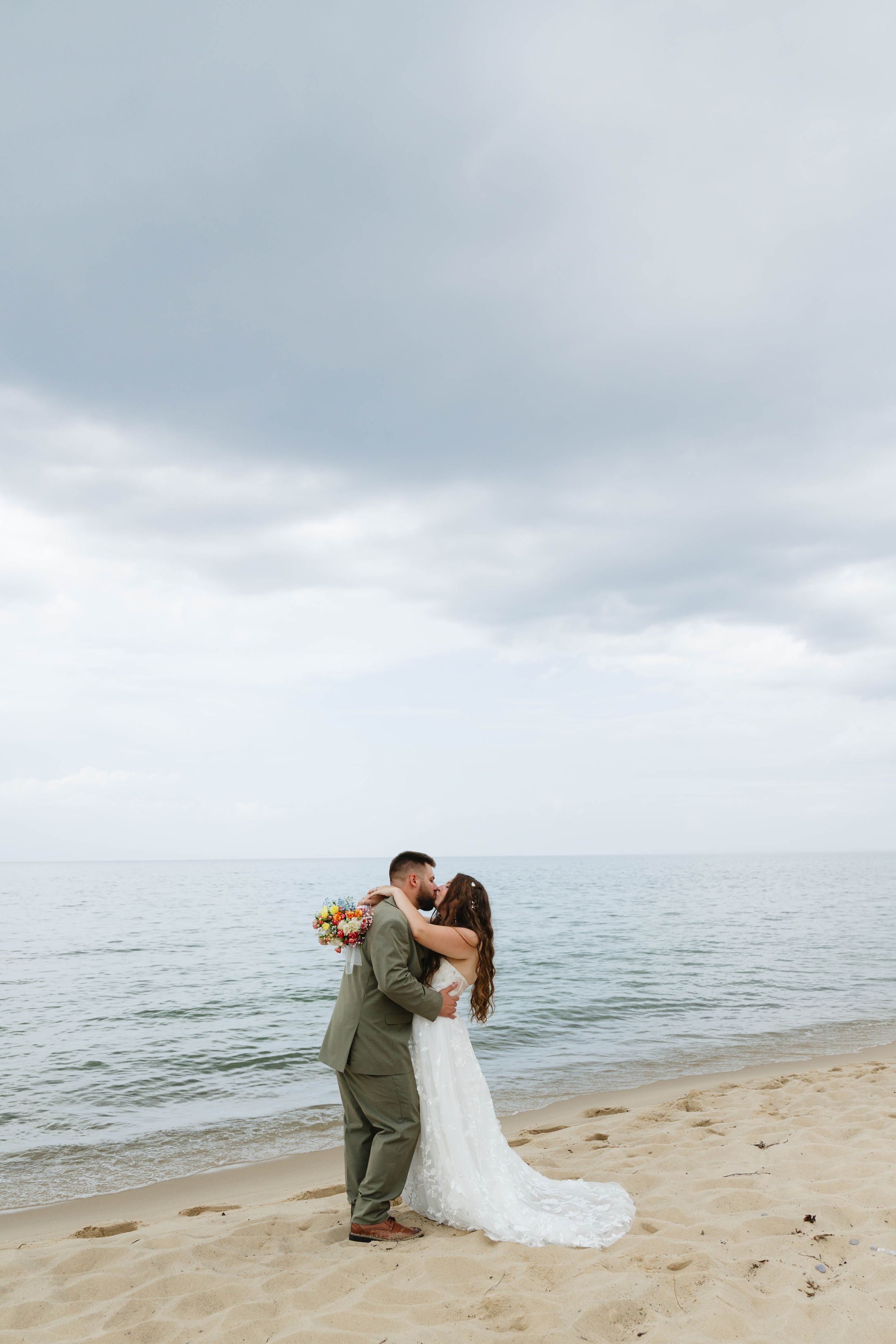 A romantic couple in wedding attire share an intimate moment while dancing on a cloudy beach.