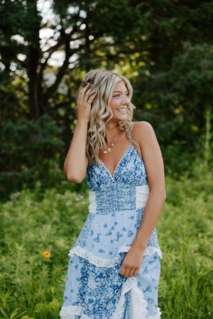 A woman in a blue and white floral dress poses outdoors in a grassy field during golden hour.