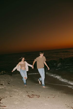 Two people run playfully along a dark beach at sunset with ocean waves in the background.