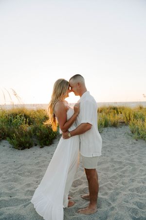 A romantic couple embraces on a sandy beach at sunset with sea oats and dunes in the background.