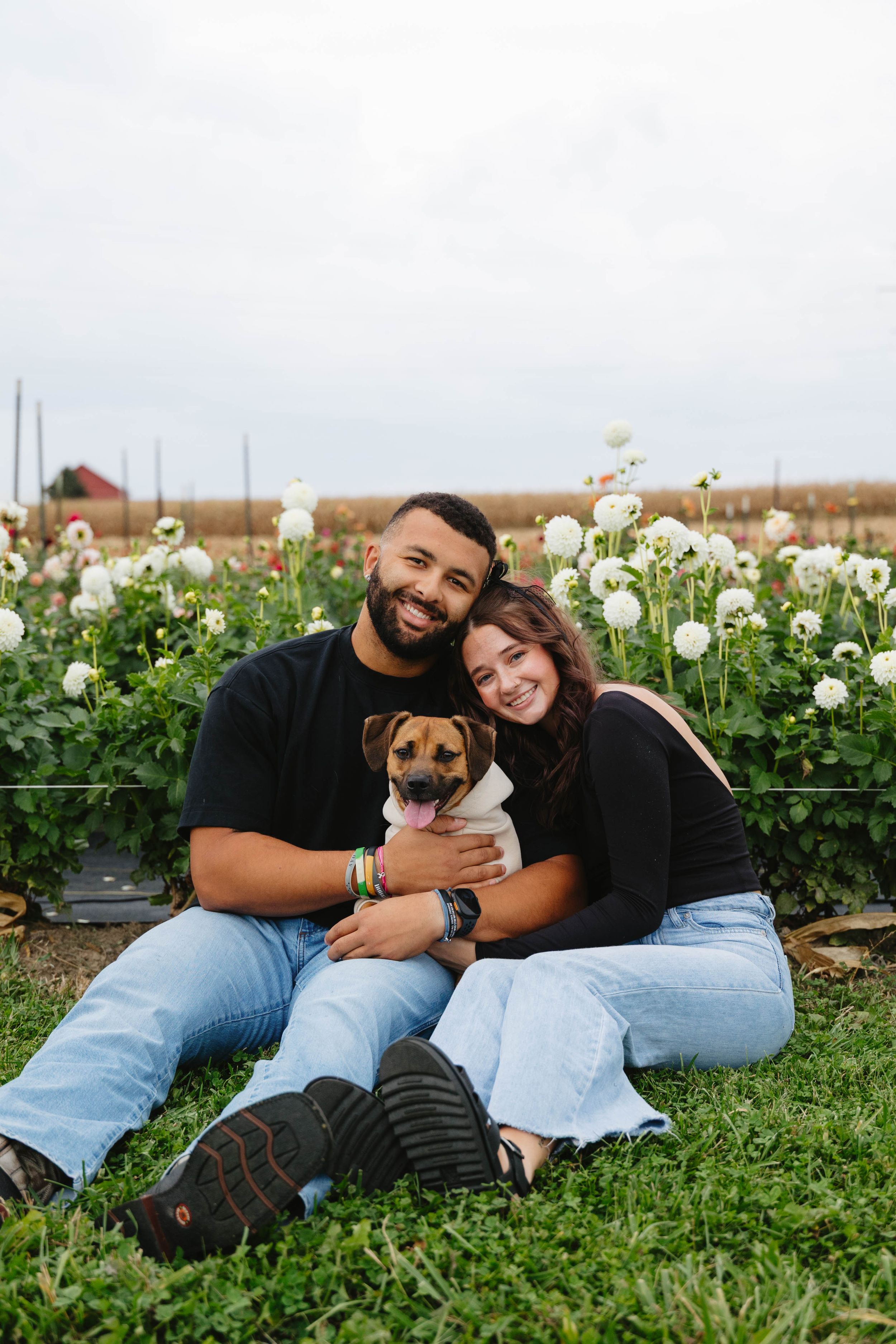 A couple sits together on grass with their small brown dog in front of white flowers blooming in a garden.