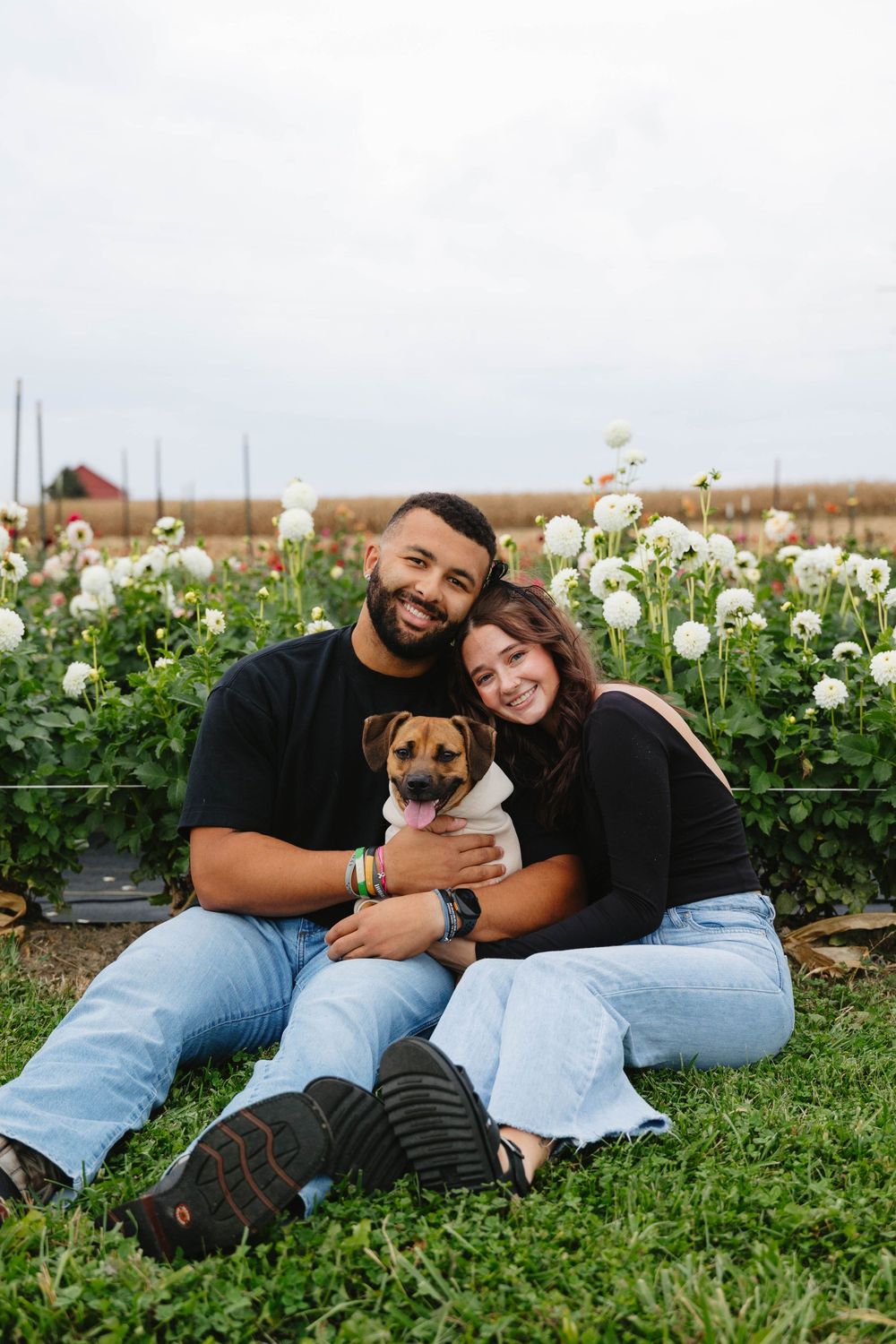 A couple sits together on grass with their small brown dog in front of white flowers blooming in a garden.