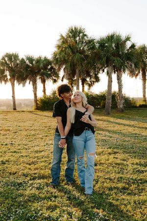 Couple embraces in a romantic sunset moment among palm trees and wildflowers in a grassy field.