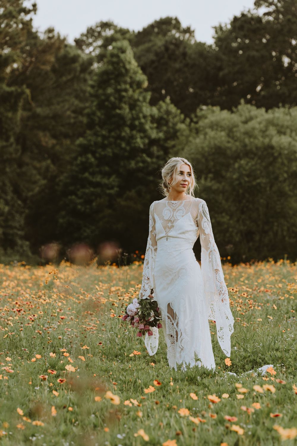 Ethereal bride in long-sleeved lace gown standing in a wildflower meadow at sunset.