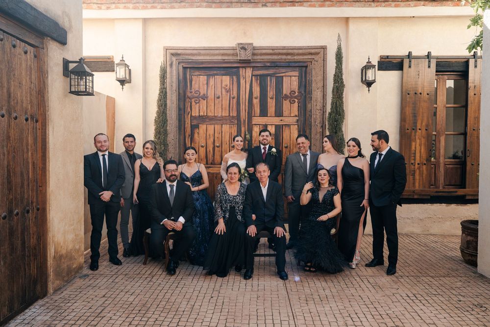 Wedding party group photo in rustic courtyard with wooden doors and stone architecture.