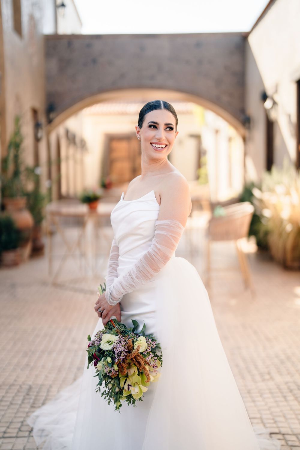 Bride twirling in elegant white gown while holding colorful bouquet in stone archway.