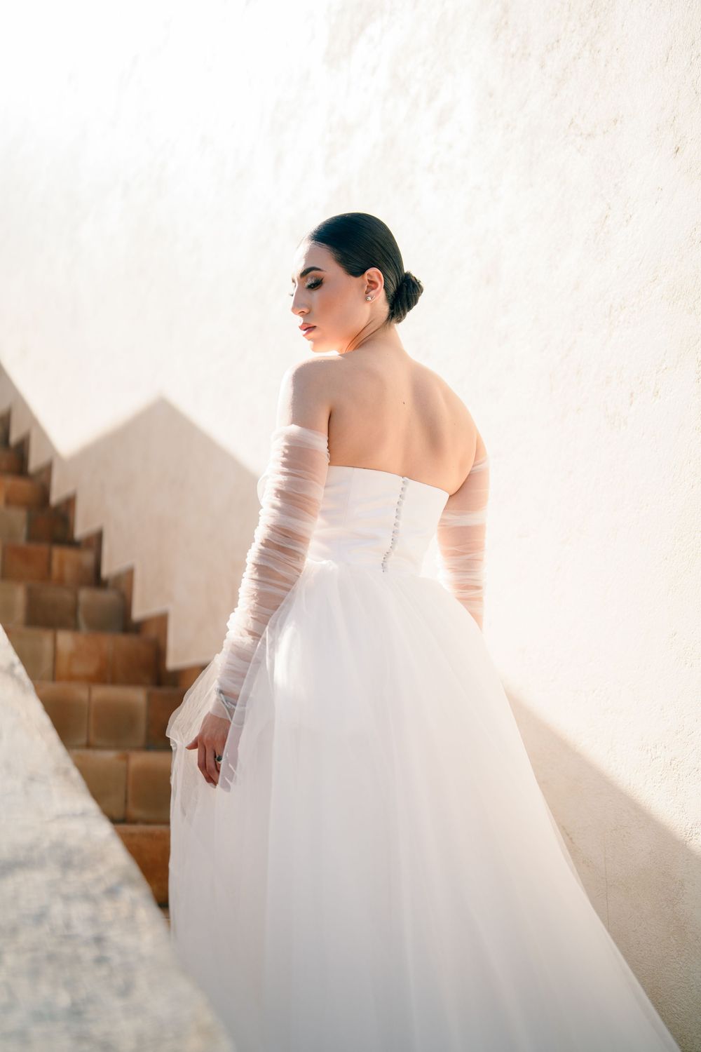 Elegant wedding dress with long sleeves and full skirt photographed in natural light against white wall.