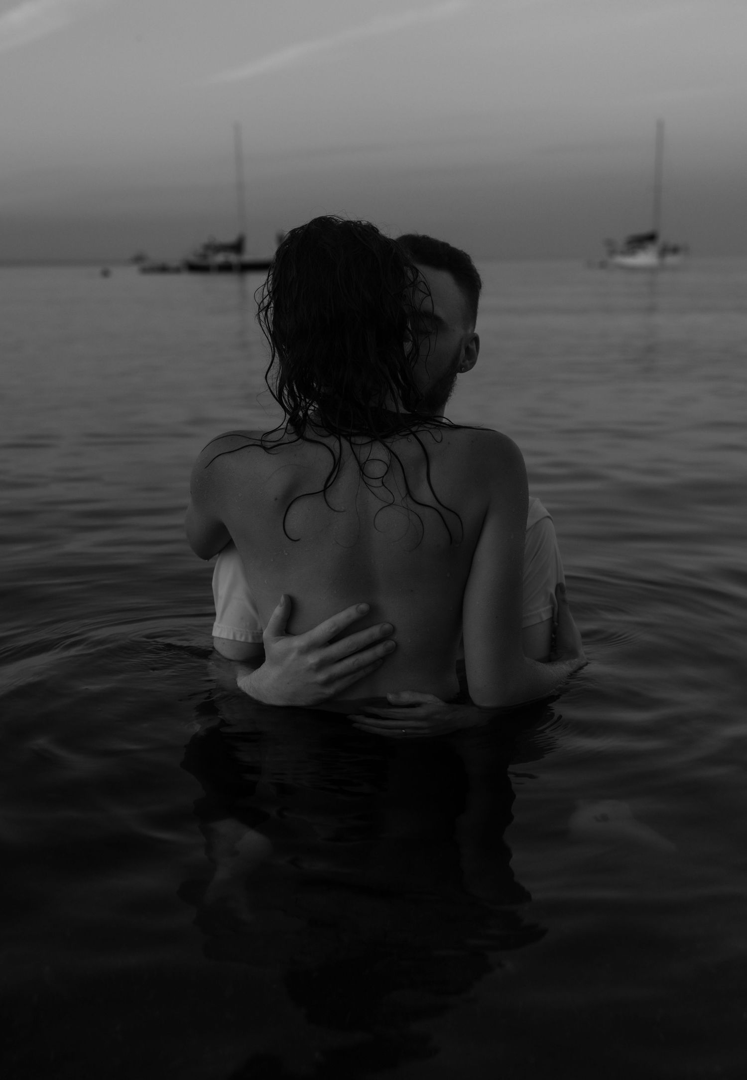 Couple embracing in dark waters with boats visible on the horizon at dusk.