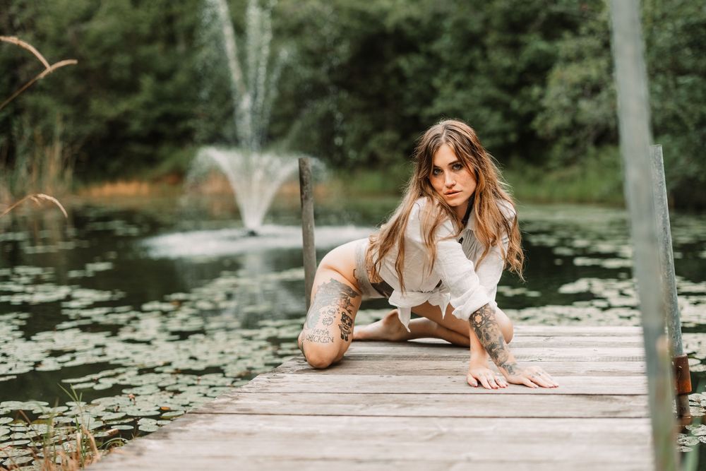 Woman in a romantic white dress lies on wooden dock near pond with lily pads during golden hour.