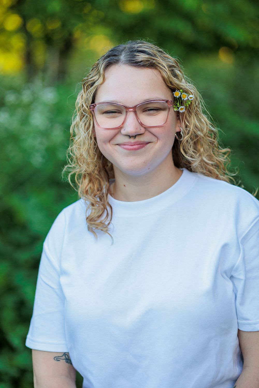Young woman with curly blonde hair, pink glasses, and white t-shirt smiling outdoors with green foliage background.