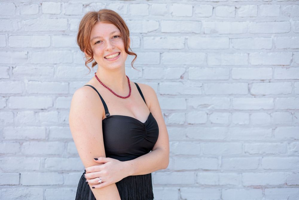 Smiling redhead woman in black dress with beaded necklace posing against white brick wall.
