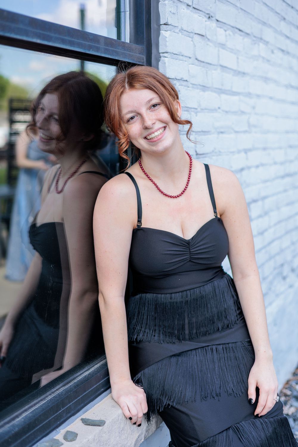 Young woman in black fringed dress and red necklace smiling, leaning against a window with her reflection visible.